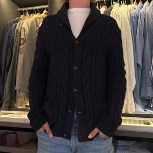 Mörkblå kofta från Polo Ralph Lauren - Säljer en mörkblå, grovstickad kofta från Polo Ralph Lauren med snyggt vågigt mönster och bruna mockadetaljer på armbågarna. Koftan har knappar framtill, två fickor och muddar