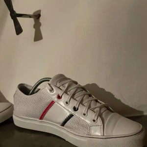 Vita Lacoste sneakers med färgdetaljer - Skriv ett Relativt PRIS!!   Snygga vita sneakers från Lacoste med perforerade sidor och coola detaljer i rött och mörkblått. Skorna har snörning och rund tå, samt en diskret Lacoste-logga på sidan. Perfekt för dig som gillar stilrena och sportiga vibes.
