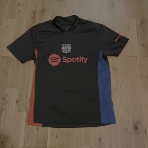 FC Barcelona Lamine Yamal tröja svart - Säljer en FC Barcelona matchtröja med Lamine Yamal #19 på ryggen. Tröjan är svart med röda och blå sidopaneler, har korta ärmar och tryckta sponsorer som Spotify och UNHCR. FCB-emblemet i svartvitt på bröstet. Tillverkad i lätt polyester, perfekt för fotboll. 