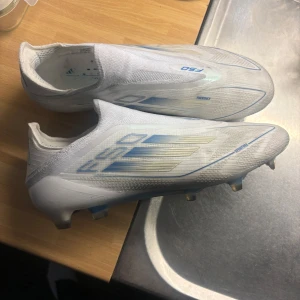 Vita Adidas F50 fotbollsskor - Säljer ett par vita Adidas F50 fotbollsskor med blå detaljer och stilren design. Skorna har låg profil, rund tå och platt sula med fasta dobbar för spel på gräs. Perfekta för dig som vill ha lätta och snabba skor på planen.