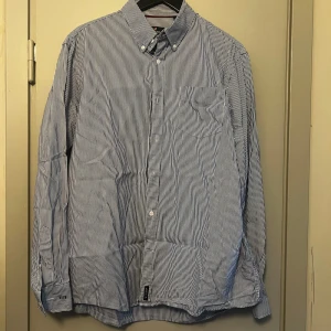 Blåvit randig skjorta från U.S. Polo Assn. - Säljer en klassisk blåvit randig skjorta från U.S. Polo Assn. i 100% bomull. Skjortan har lång ärm, knappar framtill och button-down krage. Perfekt för en stilren och avslappnad look.