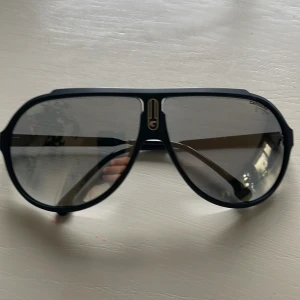 Svarta Carrera pilotbågar solglasögon - Snygga svarta solglasögon från Carrera med klassisk pilotform och gråtonade linser. Ramen är i matt svart plast med guldiga Carrera-detaljer på både front och skalmar. Perfekt accessoar för en cool och sportig look.