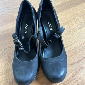 Svarta klackskor med rem från Ecco - Säljer ett par svarta klackskor från Ecco med rund tå och justerbar rem över foten. Skorna har en stabil klack och är tillverkade i skinn, vilket ger en klassisk och stilren look. Perfekta för dig som vill ha både komfort och stil.