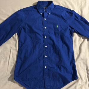 Blå skjorta från Ralph Lauren - Snygg blå skjorta från Ralph Lauren i custom fit-modell. Skjortan har klassisk krage med knappar, broderad på bröstet. Tillverkad i bomull och stängs med vita knappar framtill.