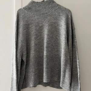 Grå stickad polotröja - Mysig grå stickad polotröja med bred ribbad krage och ribbade muddar. Tröjan har en loose passform och långa ärmar, perfekt för kyliga dagar. Enkel att styla med jeans eller kjol för en avslappnad look.