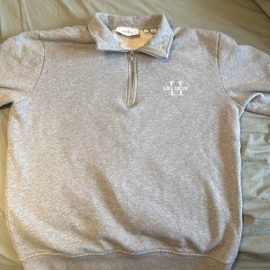 Grå halvzip sweatshirt från Les Deux - Säljer en stilren grå sweatshirt från Les Deux med halv dragkedja och ståkrage. Tröjan har logga tryckt på bröstet och är tillverkad i mjukt material. Perfekt för en avslappnad och snygg look.