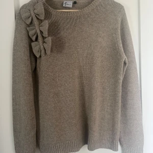 Beige stickad tröja med rosetter  - Vacker beige stickad tröja med rosetter längs axeln!!Tröjan har rund halsringning och långa ärmar. Storlek M men passar även S då den är ganska strechig. Använd 1 gång och fint skick💖
