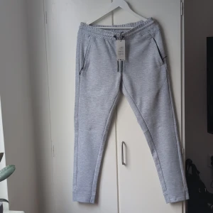 Grå mjukisbyxor Jack & Jones L - Snygga ljusgrå mjukisbyxor från Jack & Jones med svarta dragsnören och dragkedjor vid fickorna. Byxorna har normal passform och är tillverkade i mjukt bomullsmaterial. Perfekta för chill dagar eller när du vill vara bekväm men ändå se fräsch ut.