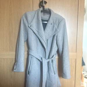 Grå kappa med bälte från Vero Moda - Snygg grå kappa från Vero Moda med bälte i midjan och dragkedjor vid fickorna. Jackan har bred krage, långa ärmar med knappar vid ärmslut och en stilren look. Perfekt för dig som vill ha en klassisk men modern vibe.