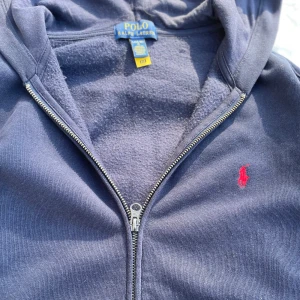 Mörkblå zip från Ralph Lauren - Säljer en snygg mörkblå hoodie från Ralph Lauren med dragkedja och huva. Den har en röd broderad logga på bröstet och praktiska fickor framtill. Perfekt för en avslappnad stil. Tröjan är i bra skick och är lite solbränd. Skriv till mig om fler bilder eller frågor💛