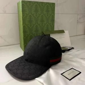 Svart Gucci Keps - Exklusiv svart keps från Gucci med diskret GG-monogram över hela kepsen. Klassisk böjd skärm och ikoniskt grönt och rött band på sidan. Tillverkad i bomull och levereras med originalpåse och box. Perfekt för dig som vill ha en lyxig streetstyle-accessoar. ÄKTA!!!!