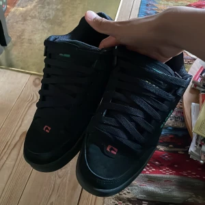 Svarta Globe sneakers med färgdetaljer - Tunga svarta sneakers från Globe med gröna, röda och gula detaljer. Skorna har bred platt sula, rund tå och tjock vaddering runt ankeln. Klassisk skate-stil med snörning och logga på sidan och hälen. Perfekta för dig som gillar streetwear och skate.