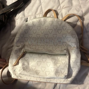 Vit ryggsäck från Valentino - Snygg vit kanske lite mer åt det beiga hållet ryggsäck från Valentino med all-over logomönster och ljusbeiga detaljer. Väskan har en stor ytterficka med dragkedja och flera fack inuti. Tillverkad i syntetmaterial med stilrena detaljer och justerbara axelband. Nypris 1645kr
