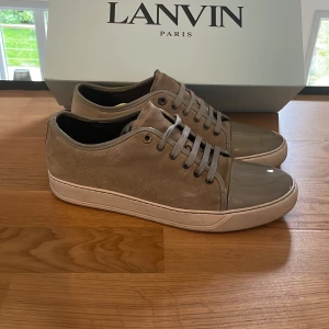 Lanvin skor - Säljer ett par stilrena sneakers från Lanvin i färgen grå och beich med grå snörning och glansig, lackad tå. Skorna har vit sula och diskret logga på sidan. Perfekta för dig som gillar en clean och lyxig look. Skorna är perfekt inför sommaren!