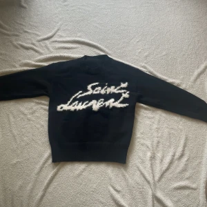 Svart  stickad tröja från Saint Laurent - Säljer en mörkblå stickad tröja från Saint Laurent med vit, suddig logotyp framtill. Tröjan har rund halsringning, långa ärmar och breda ribbade muddar vid ärmslut och nederkant. Perfekt för dig som gillar exklusiva och unika plagg.