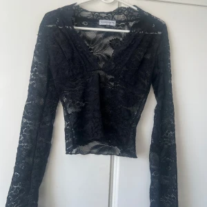 Svart spetsblus från Venderby's - Säljer en svart, transparent spetsblus från Venderby's i storlek XS. Blusen har långa ärmar, v-ringning och ett blommigt spetsmönster.