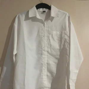 Vit klassisk skjorta med bröstficka - Vit skjorta med klassisk krage, lång ärm och bröstficka. Skjortan har knappar hela vägen framtill och är tillverkad i bomull. Perfekt för en clean och stilren look. Helt oanvänd 