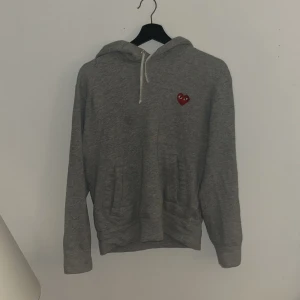 Grå hoodie Comme des Garçons Play S - Grå hoodie från Comme des Garçons Play i 100% bomull. Klassisk modell med huva, snörning och känguruficka. På bröstet sitter det ikoniska röda hjärtat med ögon. Perfekt för dig som gillar streetwear och stilrena plagg. Inköpt på Nk för 2600 använd i cirka 1 år mint skick inga skador