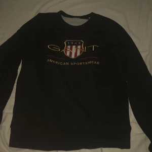 Svart sweatshirt från Gant - Svart sweatshirt från Gant med broderad logga och sköld på bröstet. Tröjan har lång ärm och klassisk passform. Snyggt broderi i gult, rött och vitt ger en sportig vibe. Perfekt för dig som gillar stilren streetwear.