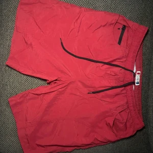 Röda badshorts från Clique, storlek L - Röda badshorts från Clique i storlek L. De har elastisk midja med svart snörning, meshfoder och praktisk ficka med dragkedja fram. Shortsen är tillverkade i lätt syntetmaterial och har ventilerande hål bak. Perfekta för stranden eller poolen!