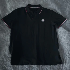 Svart pikétröja från Moncler - Säljer en svart pikétröja från Moncler med klassisk krage och knäppning framtill. Tröjan har Moncler-logga broderad på bröstet och detaljer i rött och vitt på krage och ärmslut. Perfekt för en stilren look.