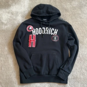 Svart hoodie från Hoodrich  - Svart hoodie från Hoodrich med vita och röda patchar och broderade detaljer framtill. Stor huva och magficka. Stora tryck och broderier på bröstet ger en streetwear-känsla.
