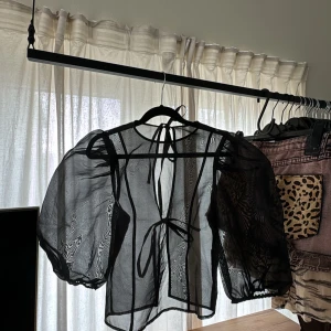 Svart mesh blus med knytband - Säljer en svart transparent blus med puffiga ärmar och knytband både i nacken och framtill. Blusen har en luftig och lätt känsla, perfekt för att styla lager på lager. Snygg detalj med de tunna banden och den genomskinliga looken.