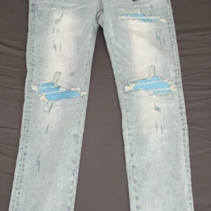 Ljusblå slitna jeans från Amiri - Ljusblå jeans från Amiri med tydliga slitningar och blå lappar under de trasiga partierna på benen. Klassisk femficksmodell med raka ben och snygga detaljer som ger en cool streetkänsla.