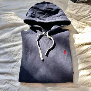 Mörkblå Polo Ralph Lauren hoodie - Säljer en mörkblå hoodie från Polo Ralph Lauren med klassisk röd broderad logga på bröstet. Tröjan har huva med vita snören och en stor magficka. Perfekt för en avslappnad stil.