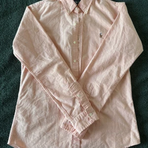 Aprikos skjorta från Ralph Lauren - Säljer en klassisk aprikos skjorta från Ralph Lauren med broderad logga på bröstet. Skjortan har lång ärm, knappar hela vägen och button-down krage. Perfekt till sommaren!