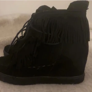 Svarta boots med fransar i mocka - Snygga svarta boots i mocka med coola fransdetaljer runt skaftet och snörning framtill. Skorna har en platt sula och ger en trendig boho-vibe, perfekta till jeans eller klänning.