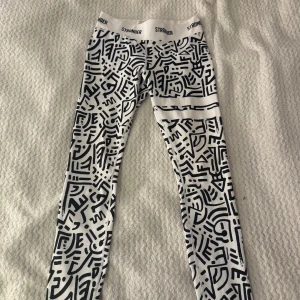 Stronger Safari leggings med grafiskt mönster - Snygga vita leggings från Stronger med svart grafiskt mönster och bred resår i midjan med logga. Modellen är högmidjad och tillverkad i stretchigt syntetmaterial, perfekta för träning eller chill. Från kollektionen Safari. Helt oanvända därför säljer jag dom för 200kr.