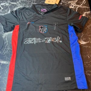 Svart FC Barcelona x Nike fotbollströja - Säljer en svart FC Barcelona fotbollströja från Nike med röda och blå sidopaneler. Tröjan har klubbmärke på bröstet, Dri-FIT-material och tryck med 'Cactus Jack' samt UNHCR-logga på ryggen. Perfekt för dig som gillar unika samarbeten.