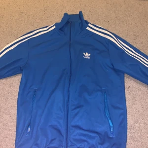 Blå Adidas track jacket med vita ränder - Säljer en klassisk blå Adidas track jacket med vita ränder längs ärmarna och dragkedja framtill. Jackan har ståkrage, två fickor med dragkedja och Adidas logga på bröstet. Perfekt för en sportig stil.