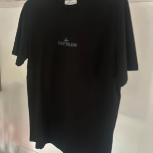 Svart t-shirt från Stone Island - Svart t-shirt från Stone Island med logga tryckt på bröstet och stort tryck på ryggen. Klassisk rund halsringning och korta ärmar. Tillverkad i mjuk bomull, perfekt för en clean och stilren look.