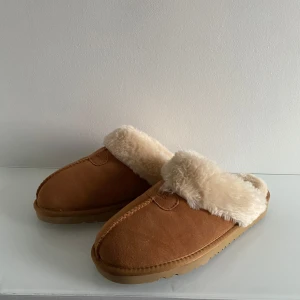 Bruna fårskinnstofflor från UGG - Mysiga bruna tofflor från UGG med mjukt, ljust fårskinnsfoder och ovandel i mocka. Perfekta för kalla dagar hemma, med en rejäl och mönstrad gummisula för extra grepp. Klassisk slip-in modell med rund tå.