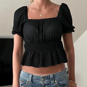 Svart croppad blus med puffärmar - Trendig svart croppad blus med fyrkantig ringning och puffiga korta ärmar. Smockad midja och volang nertill ger en snygg siluett. Perfekt till jeans eller kjol för en cool look.