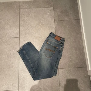 Blå slitna jeans från Nudie Jeans Co - Säljer ett par blå jeans från Nudie Jeans Co med slitningar och hål på framsidan. Jeansen har klassisk femficksdesign, orangea sömmar och knappgylf. Perfekta för en avslappnad och trendig look.