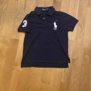 Marinblå pikétröja Polo Ralph Lauren - Snygg marinblå pikétröja från Polo Ralph Lauren i bomull, custom fit. Ikonisk vit polospelare broderad på bröstet och stor vit siffra 3 på ärmen. Klassisk krage med knappar och korta ärmar. Perfekt för en clean och sportig look.