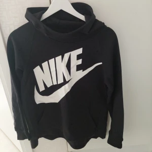 Svart Nike hoodie med stor logga - Svart hoodie från Nike med stor vit logga och swoosh över bröstet. Tröjan har huva, långa ärmar och en klassisk känguruficka framtill. Perfekt för dig som gillar sportig och avslappnad stil.                                     Från rök och djurfritt hem.                   Bilderna missvisar lite  och den är svart och ör knappt använd så den är I mycket fint skick.