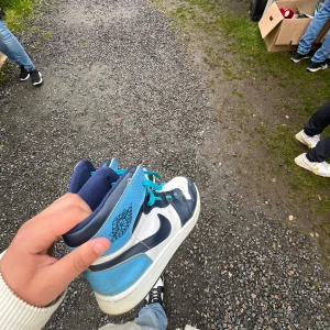 Nike Air Jordan 1 blå och vita sneakers - Säljer ett par Nike Air Jordan 1 sneakers i blått, vitt och marinblått med coola turkosa skosnören. Skorna har högt skaft, klassisk Jordan-logga på sidan och är tillverkade i skinn. Perfekta för dig som vill sticka ut med en ikonisk streetstyle. Sälja för att den e gammal