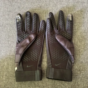 Nike handskar - Svarta Nike handskar med tryckt Nike-mönster över hela ovansidan. Insidan har greppvänliga detaljer och fingertopparna är touch-anpassade. Perfekta för träning utomhus när det är kallt. Snygg, sportig look och smidig passform.