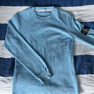 Ljusblå långärmad tröja Calvin Klein - Snygg ljusblå långärmad tröja från Calvin Klein Jeans med diskret struktur i tyget och CK-logga på ena ärmen. Tröjan har rund halsringning och ribbade muddar för en clean look. Perfekt för dig som gillar stilrena basplagg.