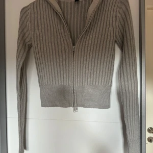 Beige ribbad kofta med dragkedja - Snygg beige ribbstickad kofta med lång ärm och dragkedja hela vägen framtill. Tröjan har en croppad passform och är perfekt för lager-på-lager. Enkel att matcha med både jeans och kjol.