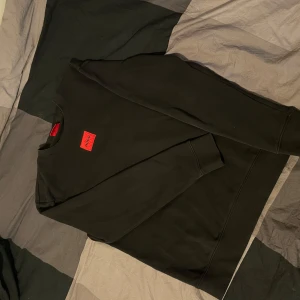Svart Hugo Boss tröja - Svart sweatshirt från Hugo Boss med röd logotyp-patch på bröstet. Klassisk rund halsringning och långa ärmar. Tröjan är i mjuk bomull och har ribbade muddar vid ärmslut och nederkant. Perfekt för en clean och stilren look.