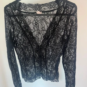 Svart spetsblus med lång ärm - Aldrig använd!! Säljer en svart blus i genombruten spets med vackert blommönster. Blusen har långa ärmar och en djup v-ringning framtill. Perfekt att styla över ett linne eller bh för en trendig look. Storlek S🤍