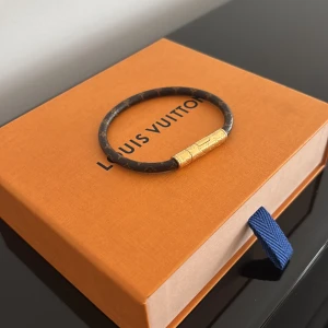 Louis Vuitton armband med gulddetalj - Säljer ett exklusivt armband från Louis Vuitton i brunt med klassiskt monogrammönster och en elegant guldfärgad låsdetalj. Perfekt accessoar för att lyfta din stil med en lyxig touch. Digitalt kvitto finns.