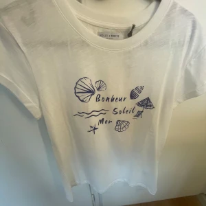 Vit t-shirt med blått tryck från Holly & White - Vit t-shirt med blått tryck av snäckor, vågor och texten 'Bonheur Soleil Mer' framtill. Klassisk rund halsringning och korta ärmar. Perfekt för en somrig look.