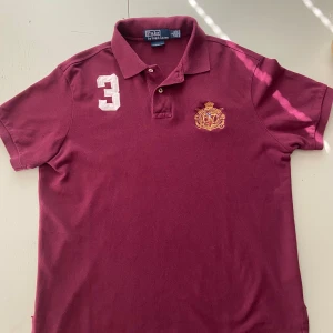 Vinröd vintage pikétröja från Polo Ralph Lauren - Säljer en vinröd pikétröja från Polo Ralph Lauren med broderad logga och siffra 3 på bröstet. Klassisk krage och knappar framtill. Är öppen för pris förslag eller byten bara att höra av er.