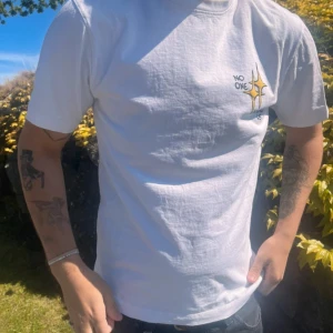 Vit t-shirt med tryck och illustration - Vit t-shirt med coolt tryck framtill där det står 'No one around us' och en stjärna. På ryggen finns en stor illustration av en tecknad figur med dödskalle som håller ett brinnande kranium. Skön passform och korta ärmar.